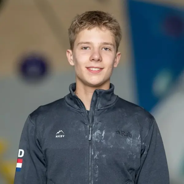 Een jongere met kort blond haar draagt een donker sportjack en lacht naar de camera. De achtergrond is wazig en bestaat uit zachte kleuren en vormen.